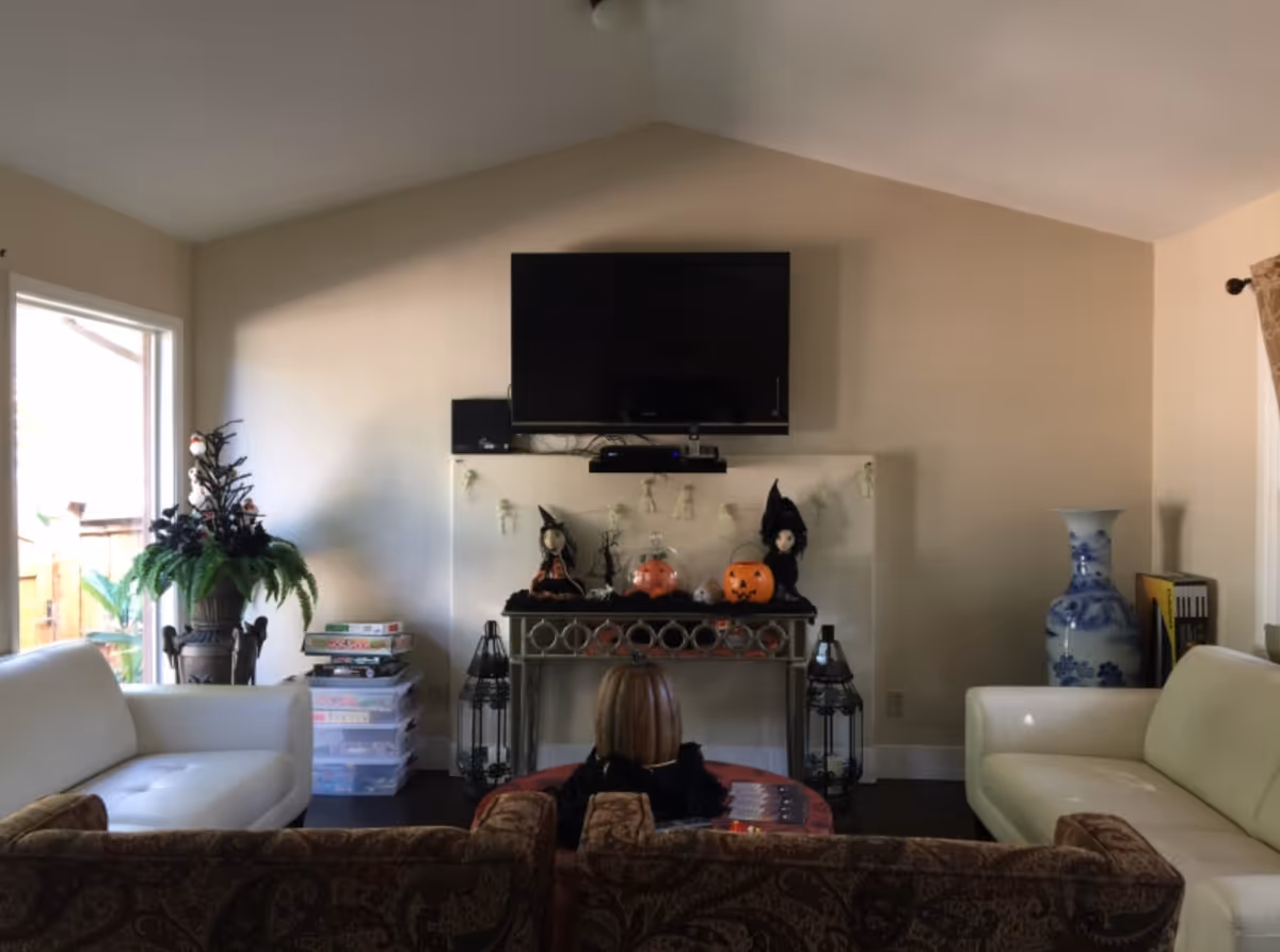A living room with a mounted flat-screen TV above a decorative console table. The table is decorated with Halloween-themed items including two witch dolls, a pumpkin, and a fishbowl with a pumpkin inside. There are two white sofas on either side and a patterned brown sofa in the foreground. A large blue and white vase is on the right side, and a potted plant is on the left near a window.