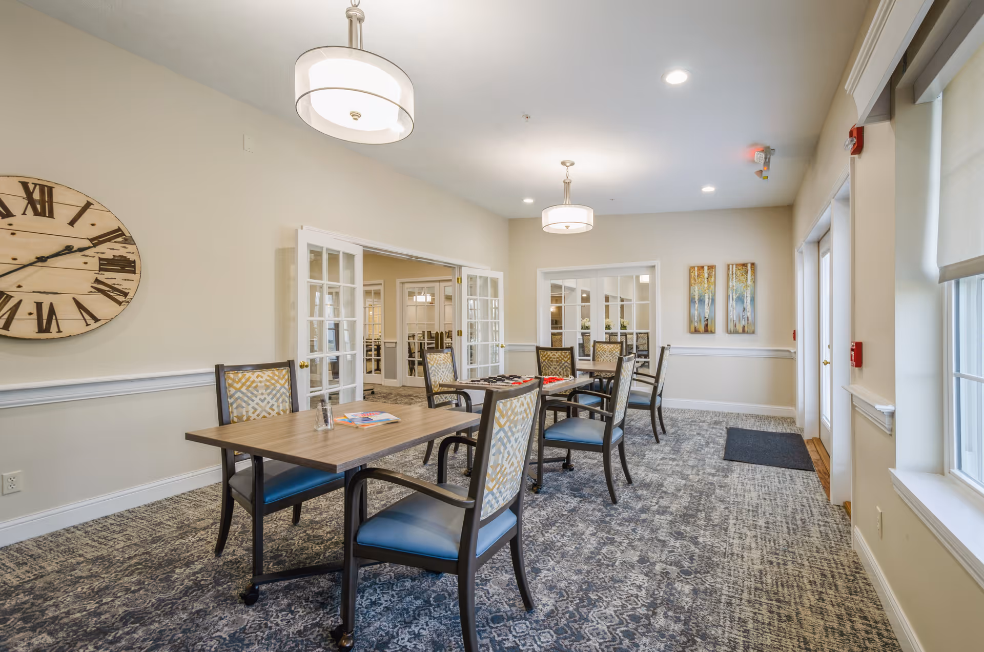 Bright communal dining area with several tables and chairs, pendant lights, a large wall clock, and windows.
