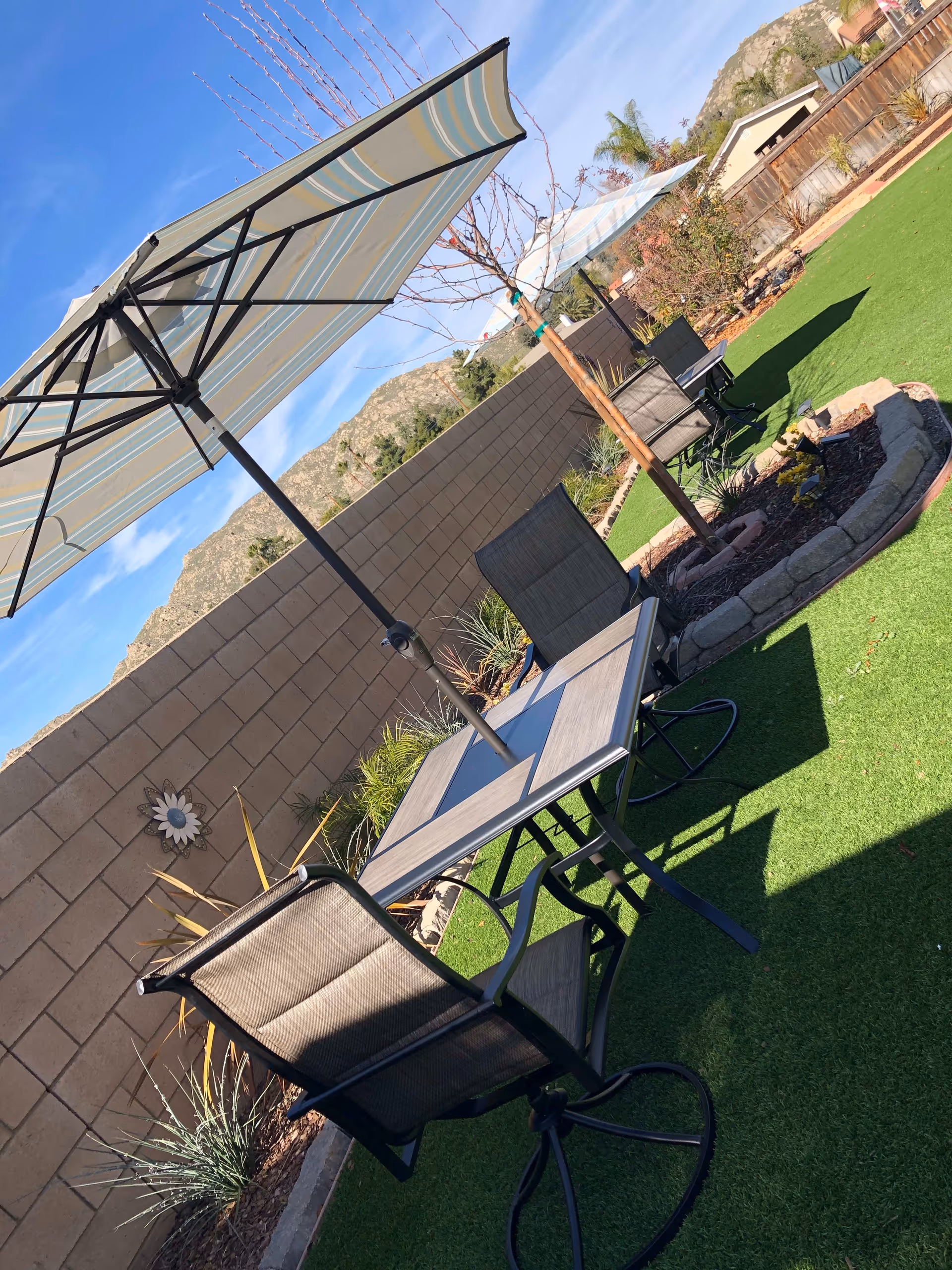 Outdoor patio area with a table and four chairs on artificial grass. Two large striped umbrellas provide shade. There is a small tree planted in a circular stone-bordered garden bed. A brick wall encloses the area, with mountains visible in the background under a clear blue sky.