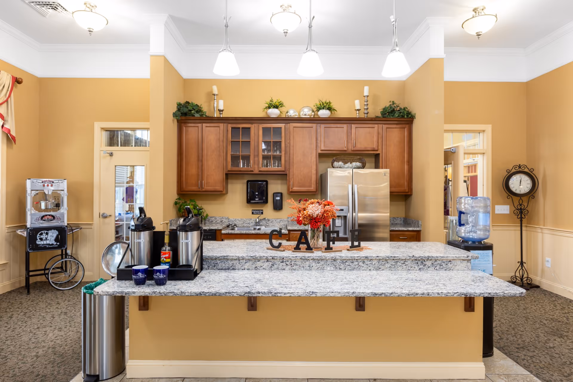 Interior view of a cafe-style kitchen with a granite island, coffee station, stainless steel refrigerator, wooden cabinets, and decorative accents.