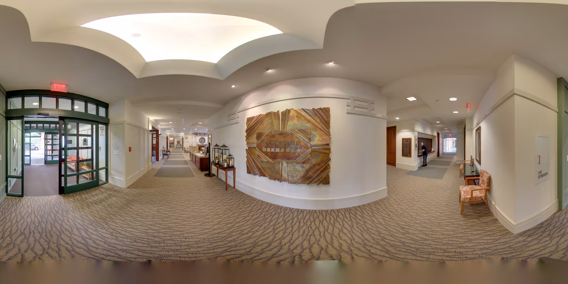 A wide-angle view of a senior living facility interior hallway with patterned carpet, cream-colored walls, and a decorative ceiling. The hallway features a large abstract wall art piece, a small table with decorative lanterns, and seating areas with chairs. There are glass double doors leading outside on the left side and a person standing in the hallway in the distance.