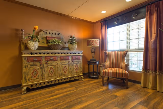 A cozy interior corner featuring a rustic wooden sideboard with decorative plants on top, a striped upholstered armchair next to a window with brown curtains, and a small round table with a lamp, all set against warm orange walls and wood flooring.