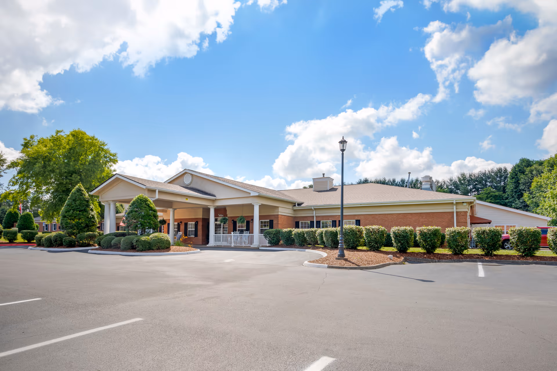 Exterior view of a single-story senior living facility building with a covered entrance, surrounded by neatly trimmed bushes and trees under a partly cloudy blue sky.