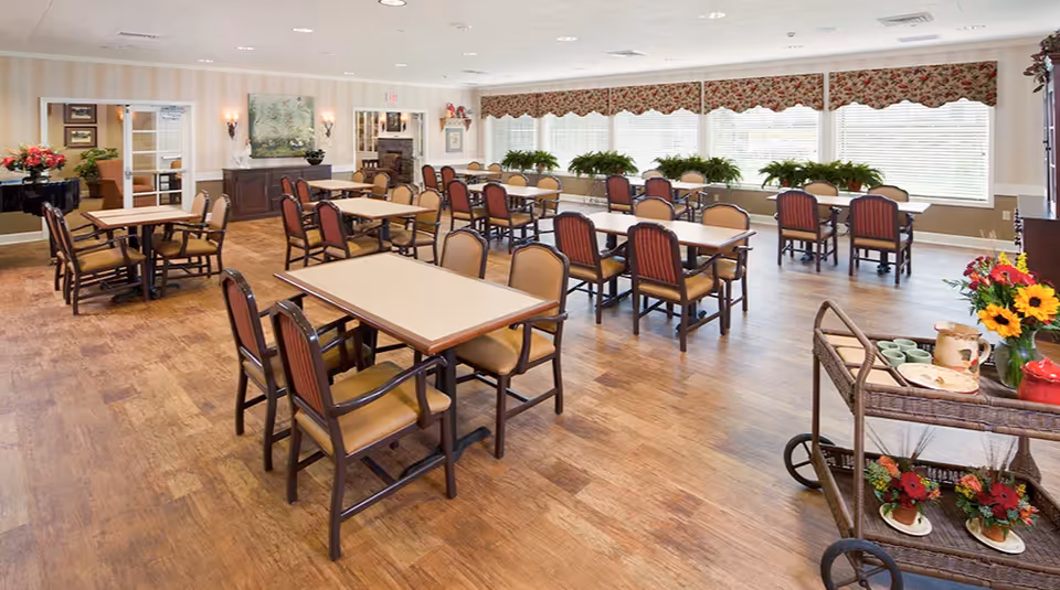 A spacious dining room with multiple tables and chairs arranged neatly on a wooden floor. Large windows with floral valances let in natural light, and several green plants are placed on the windowsills. A serving cart with cups, plates, and flower arrangements is visible in the foreground. The room has light-colored walls with decorative elements and a cozy, inviting atmosphere.