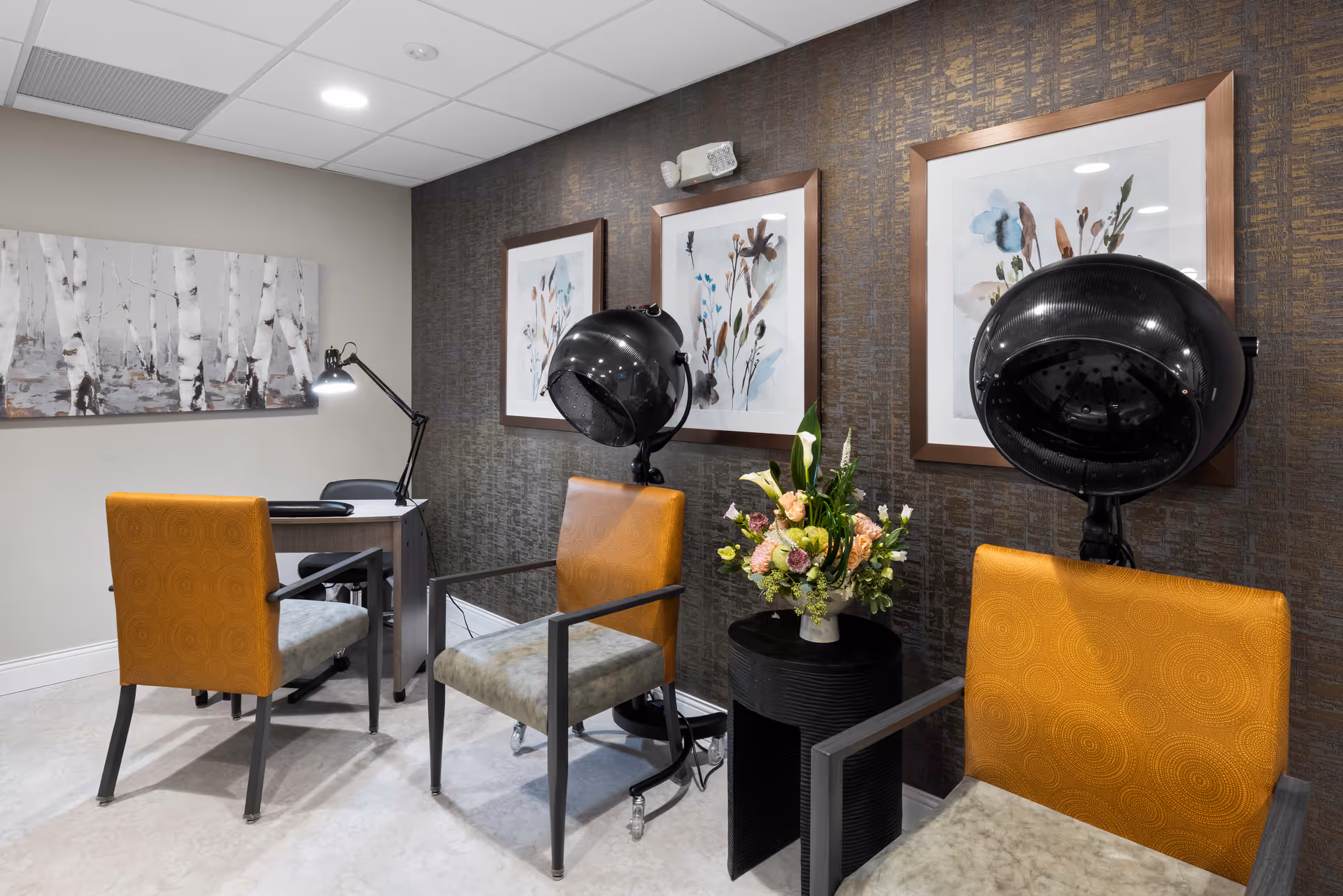 Salon-style sitting area with three chairs and hooded hair dryers, framed artwork on a textured wall, a small side table with flowers, and a desk with a lamp.
