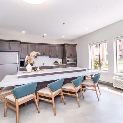 Modern kitchen with dark wood cabinets, stainless steel refrigerator and oven, a long white island countertop with five wooden chairs featuring teal cushions, and large windows letting in natural light.
