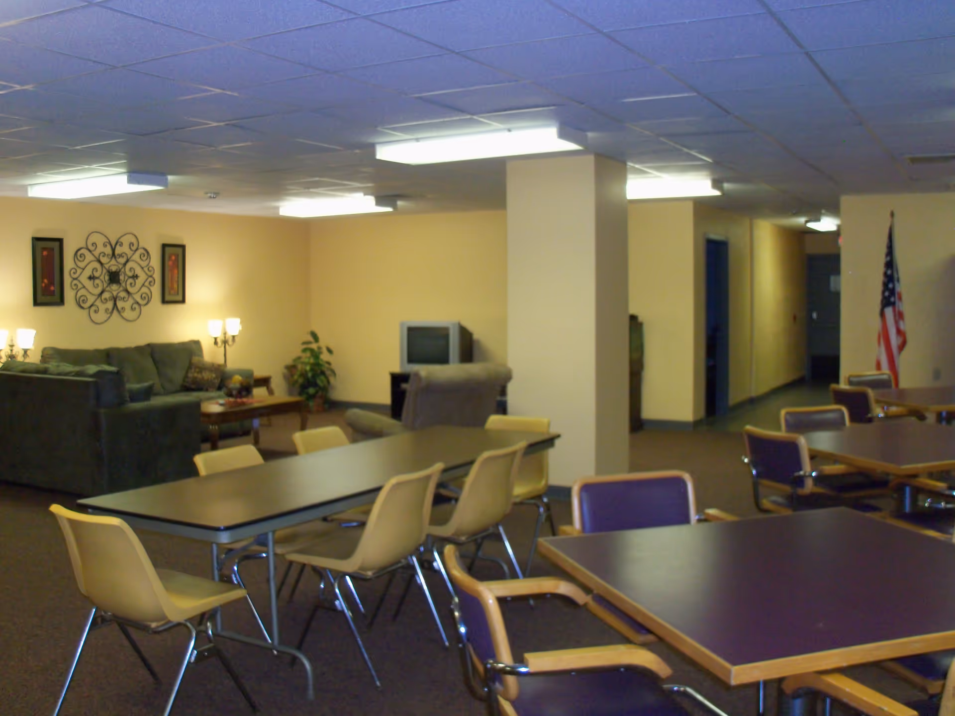 Communal activity room with multiple tables and chairs, a seating area with couches and a TV, and an American flag in the background.