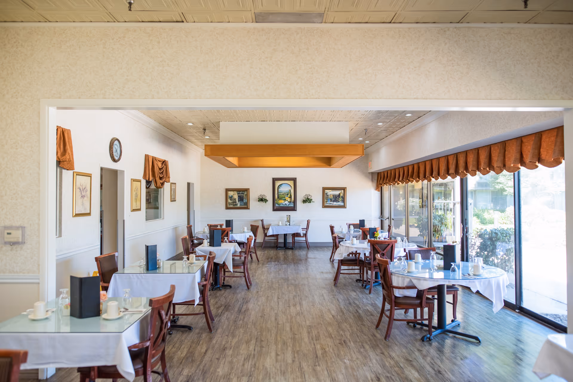 Bright dining room with multiple set tables, wooden chairs, and large windows along one side.