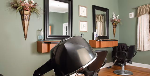 Small salon-style room with a black hooded hair dryer, two styling chairs, wall mirrors and floral wall sconces.