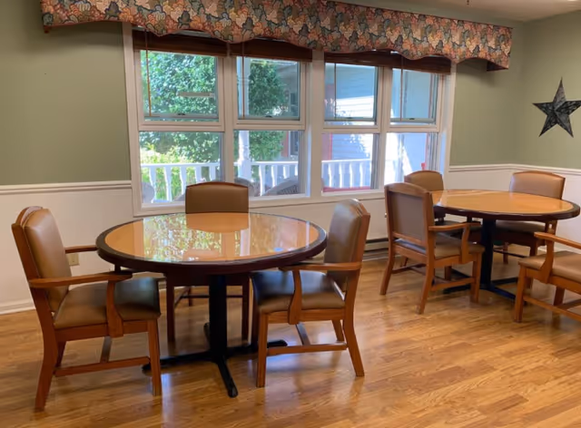 A room with two round wooden tables, each surrounded by four brown cushioned chairs. The room has light green walls with white wainscoting, a large window with a floral valance, and a wooden floor. Outside the window, greenery and part of a porch railing are visible.
