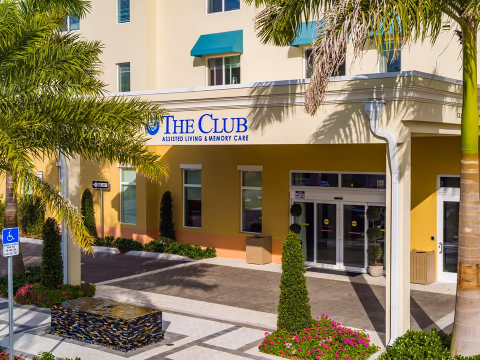 Entrance of The Club at Boynton Beach assisted living and memory care facility with a covered driveway, palm trees, decorative plants, and a wheelchair accessible parking sign.