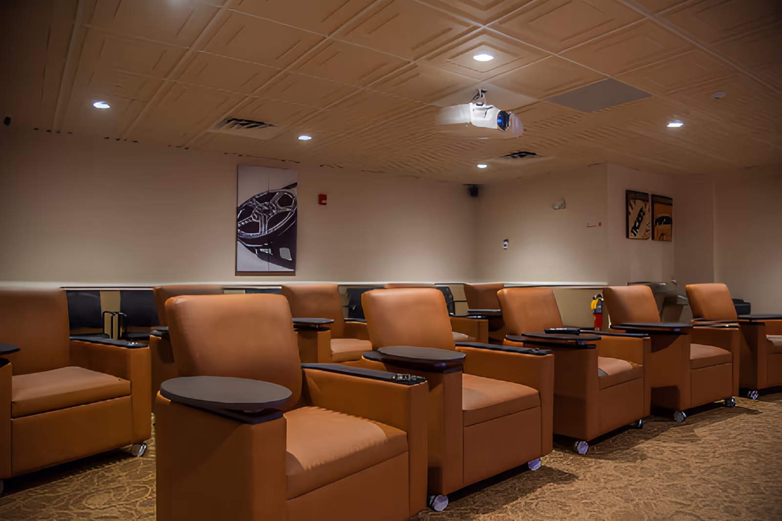 A room with multiple brown cushioned chairs arranged in rows, each with a small attached table. The ceiling has recessed lighting and a projector mounted. The walls are beige with some framed artwork and a fire extinguisher visible in the corner.