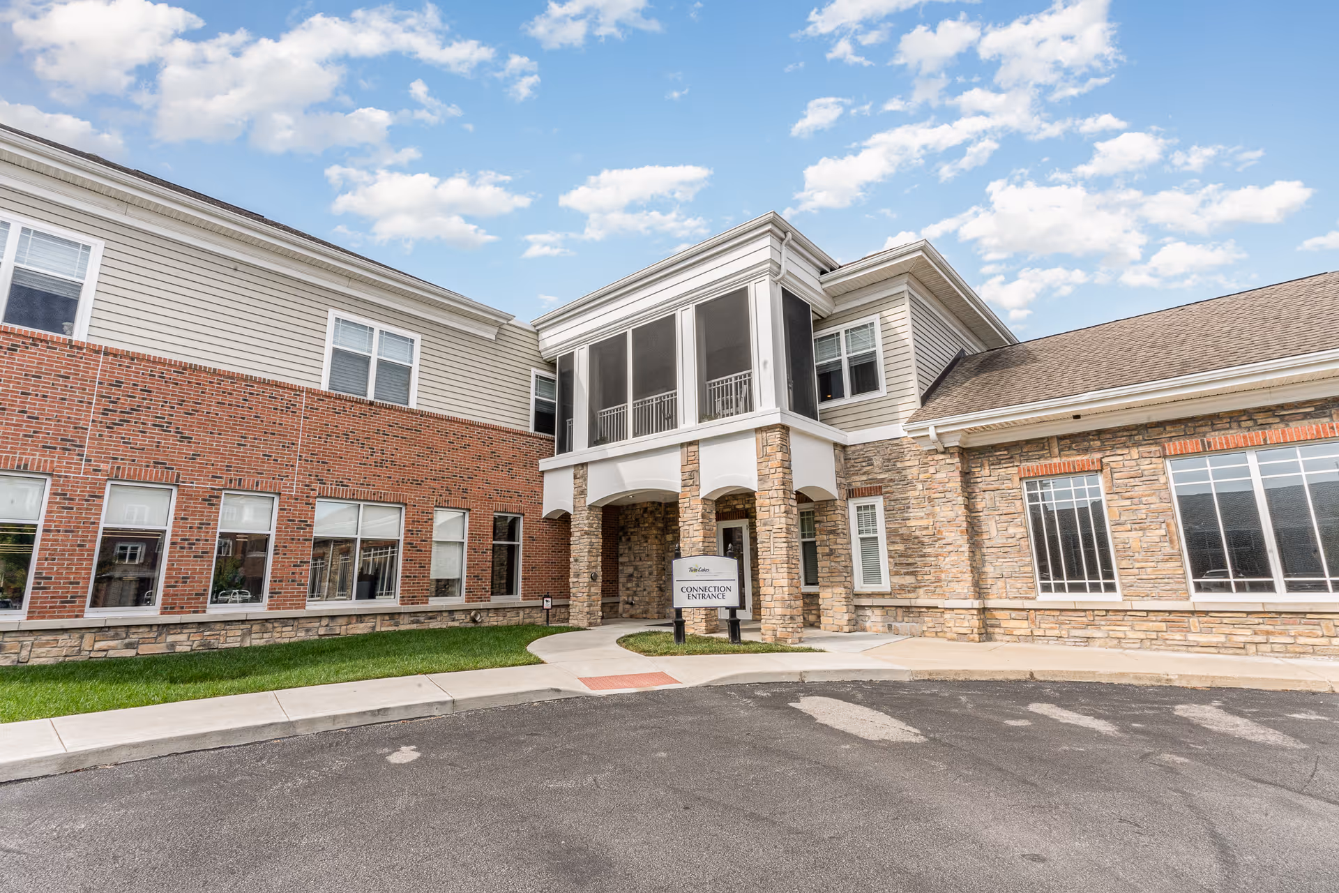 Front entrance of a brick-and-stone senior living building with a covered two-story porch, windows, and a small sign by the walkway under a blue sky.