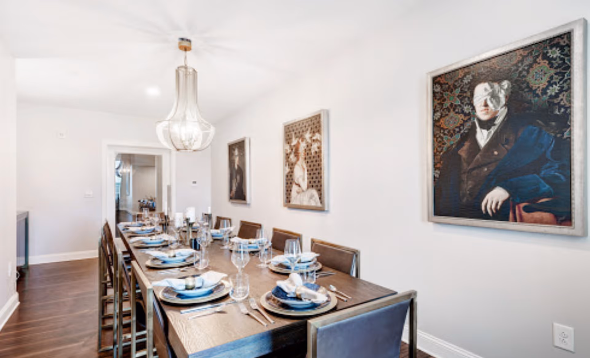 A modern dining room with a long wooden table set for eight people. Each place setting includes a plate, napkin, and glassware. The room features a contemporary light fixture hanging from the ceiling and three framed artworks on the walls. The floor is dark wood, and the walls are painted white.