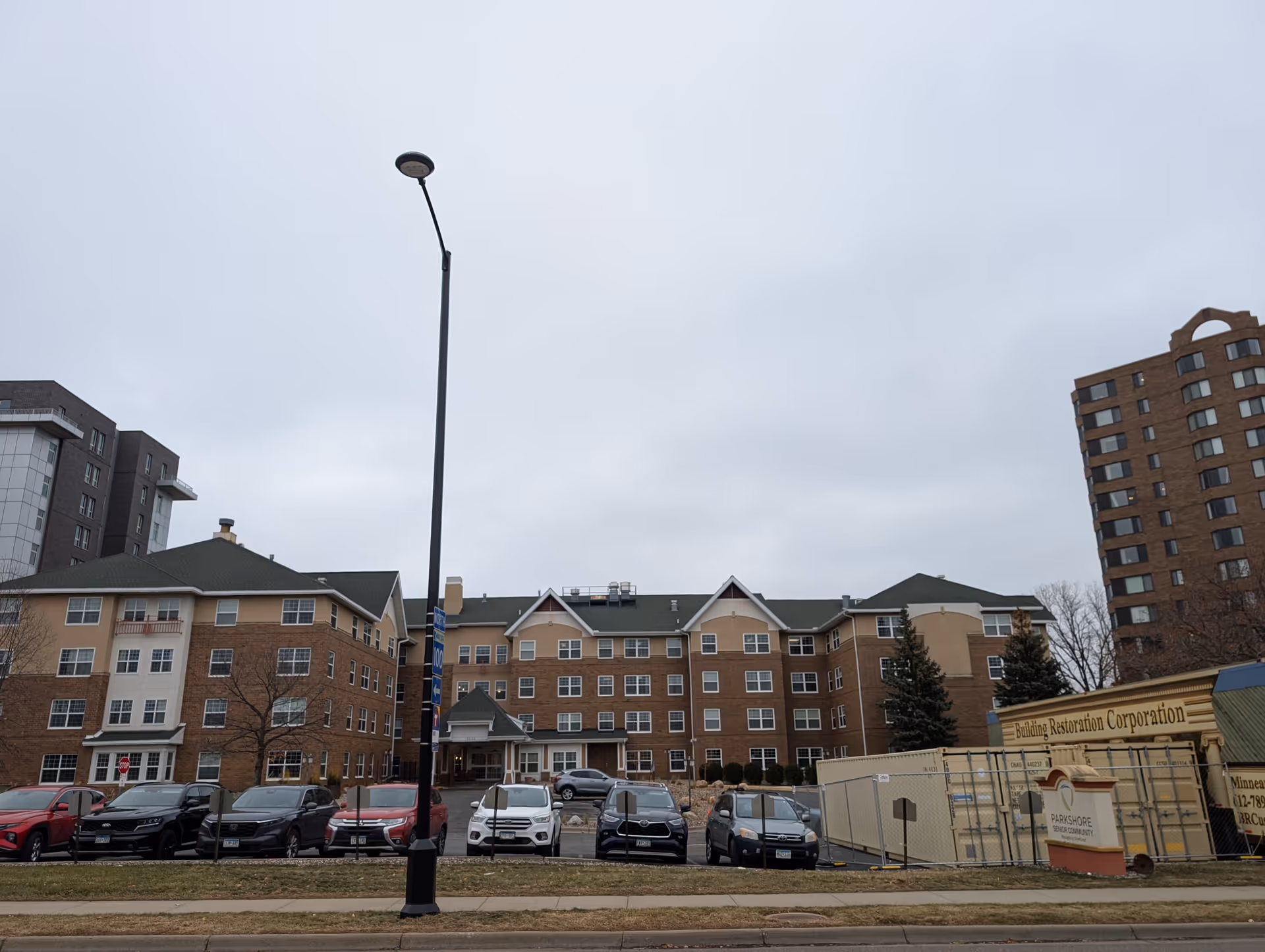 Exterior view of Parkshore Senior Campus, a multi-story brick and beige building with multiple windows. Several cars are parked in front of the building along the street. A streetlamp is visible in the foreground. There is a construction area with fencing and a sign for Building Restoration Corporation on the right side of the image. The sky is overcast.