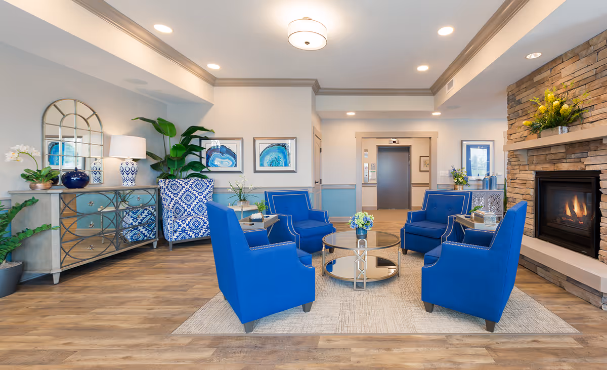Bright communal lounge with blue armchairs arranged around a glass coffee table, a stone fireplace, decorative sideboard and an elevator in the background.