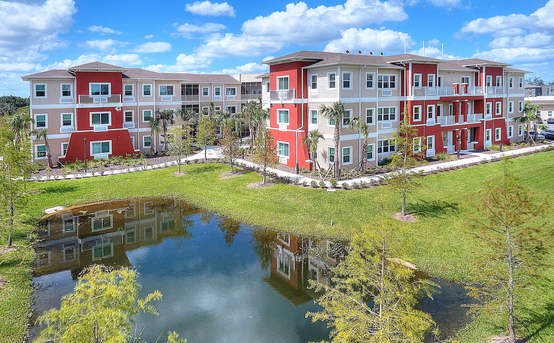 A three-story senior living facility building with red and beige exterior walls, surrounded by landscaped greenery and palm trees. In front of the building is a small pond reflecting the structure and the blue sky with scattered clouds above.