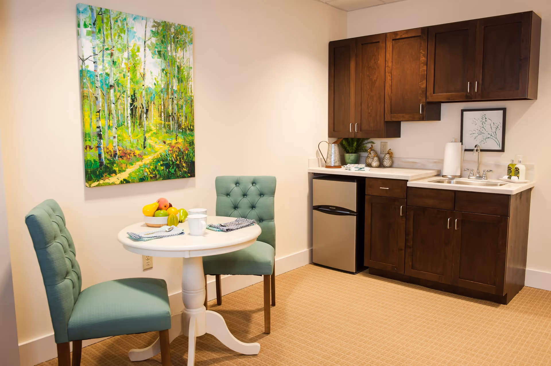 A small kitchenette area with dark wooden cabinets, a stainless steel mini refrigerator, a sink, and countertop. Next to the kitchenette is a round white table with two teal upholstered chairs. On the table are two cups, napkins, and a bowl of assorted fruit. A colorful painting of a forest scene hangs on the wall above the table.