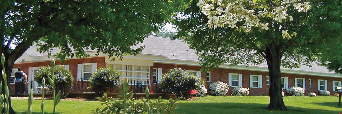 Single-story brick building with multiple windows and a white roof, surrounded by green grass, bushes, and large leafy trees providing shade.