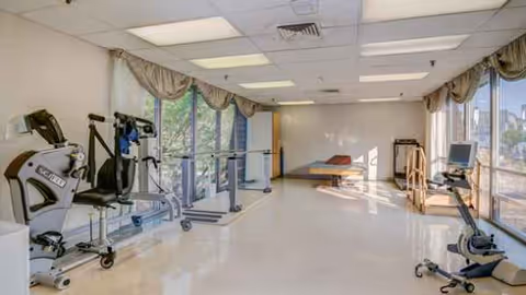 Bright rehabilitation gym with exercise machines along a wall and large windows.