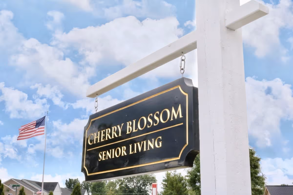 A black and gold hanging sign that reads 'CHERRY BLOSSOM SENIOR LIVING' mounted on a white post with a partly cloudy blue sky and an American flag in the background.