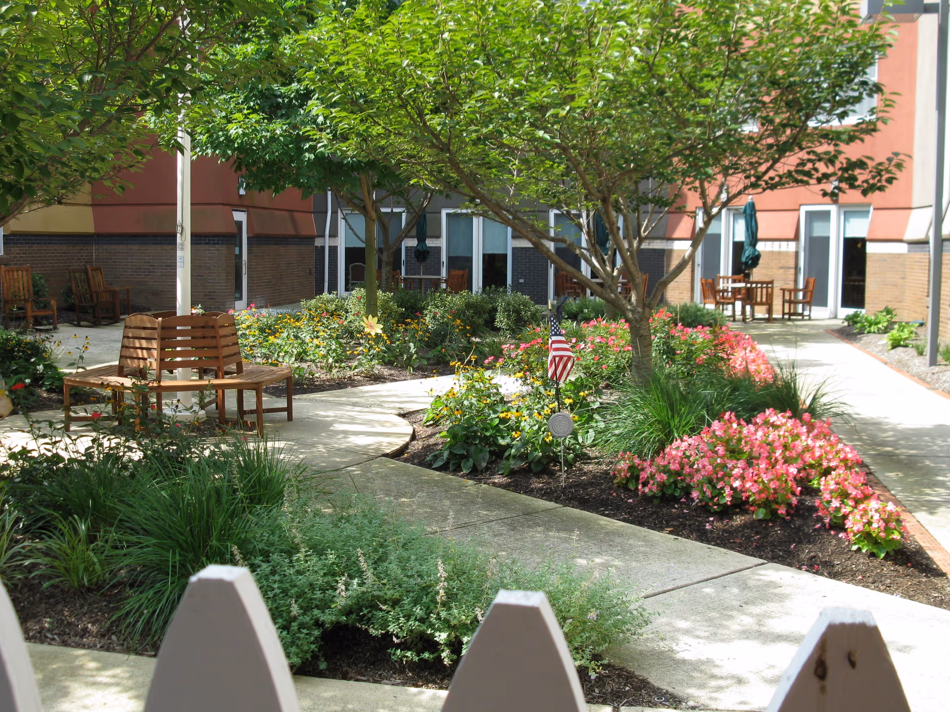 Outdoor courtyard garden area with paved walkways, green trees, colorful flower beds, wooden benches, and patio tables with chairs. The courtyard is surrounded by a building with windows and doors.