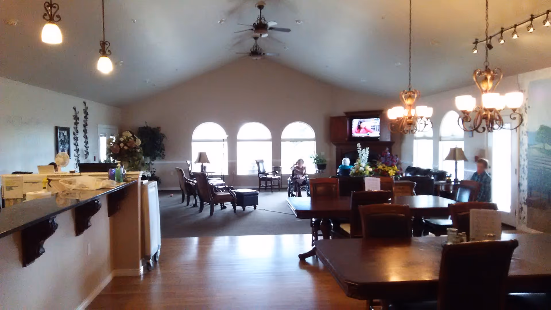 Interior view of a senior living facility common area with wooden dining tables and chairs in the foreground, a sitting area with armchairs and a wheelchair near arched windows in the background, ceiling fans, hanging light fixtures, and a television mounted above a wooden cabinet.