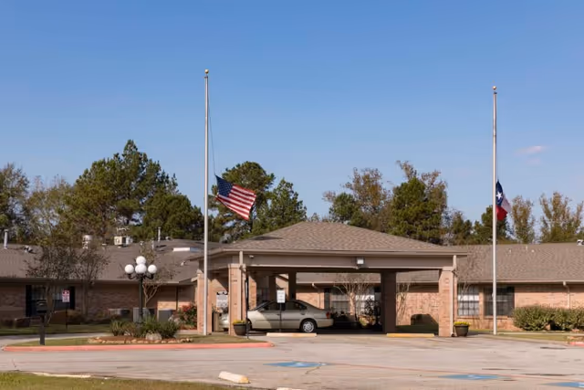Exterior view of Southland Rehabilitation And Healthcare Center showing a single-story brick building with a covered entrance. Two flagpoles with the American flag and Texas state flag flying at half-mast are visible in front of the building. There are trees and shrubs around the building and a car parked under the covered entrance.