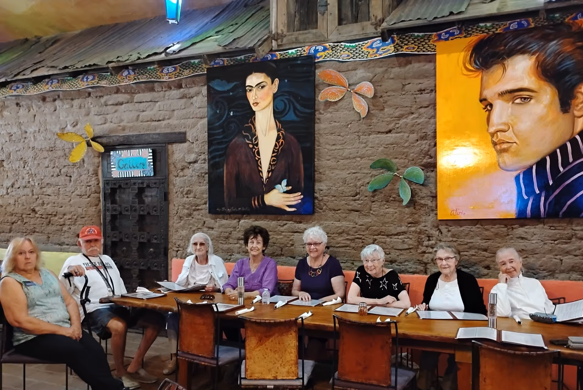 A group of people seated along a long wooden table in a colorful, art-adorned dining room.