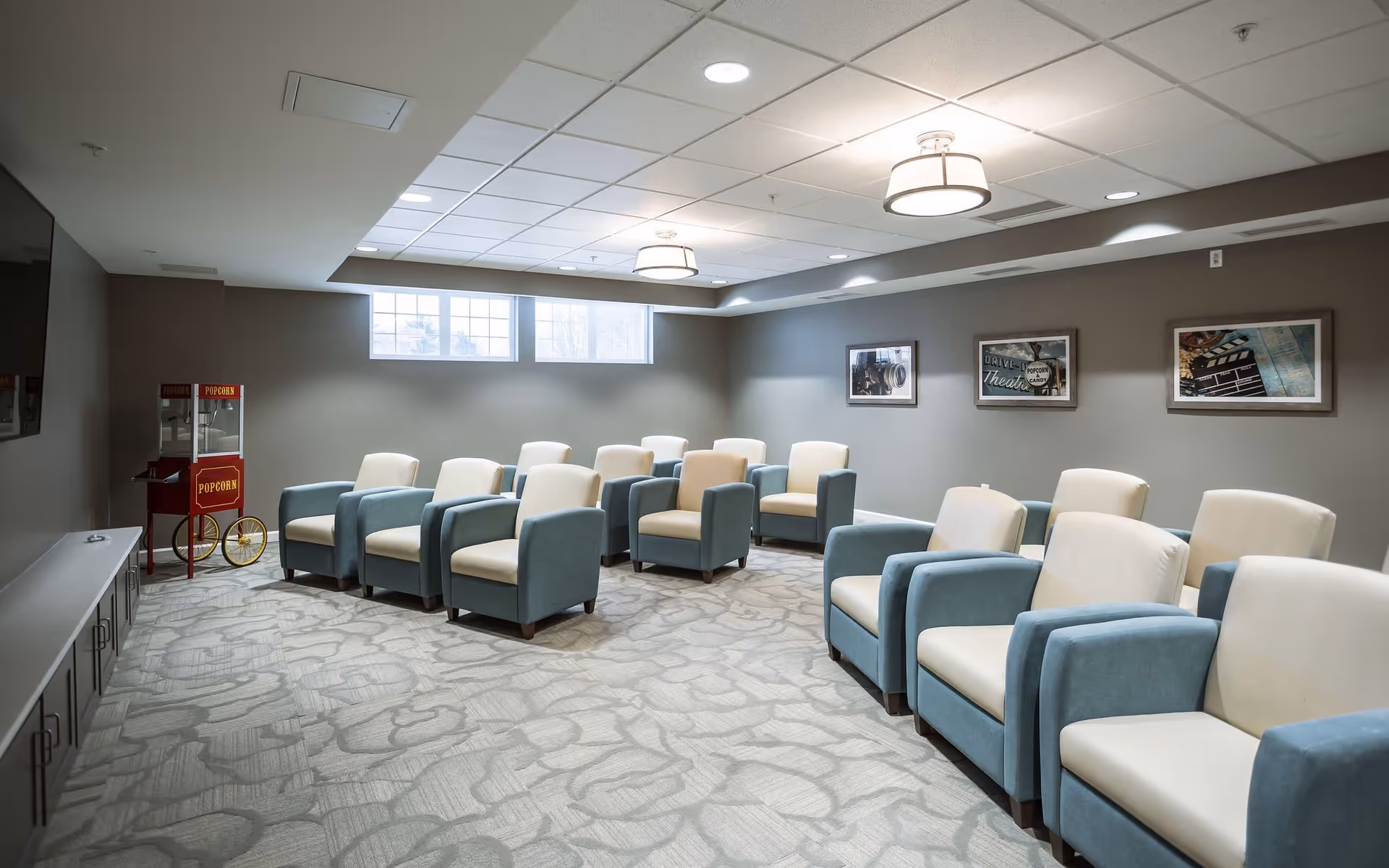 A cozy movie theater room with two rows of comfortable blue and beige armchairs facing a large flat-screen TV mounted on the wall. There is a vintage-style popcorn machine in the corner near a window letting in natural light. The walls are decorated with framed pictures related to movies and cinema.