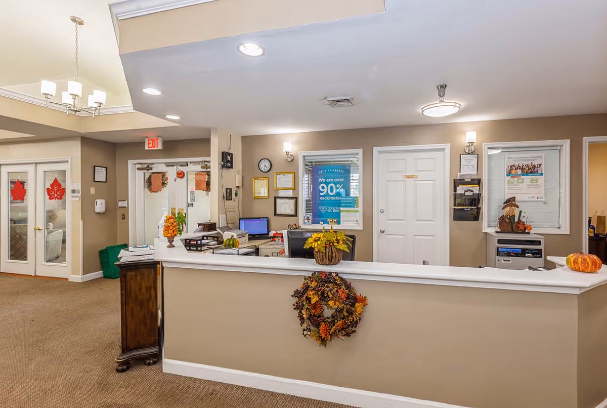 Reception area of a senior living facility with a front desk decorated with autumn-themed wreaths and small pumpkins. Behind the desk are office supplies, a computer, and informational posters on the wall. The area is well-lit with ceiling lights and has beige walls and carpeted floors. Double glass doors with red maple leaf decals are visible to the left.