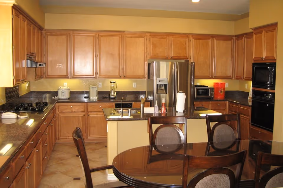 Spacious kitchen with wooden cabinets, a stainless steel refrigerator, a central island with sink, and a dining table in the foreground.
