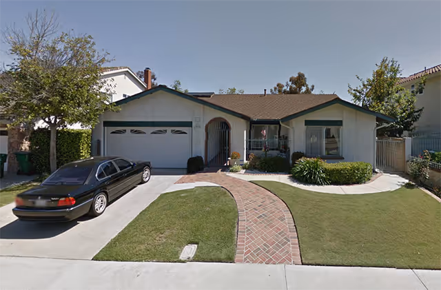 Single-story residential building with a two-car garage, a brick walkway leading to the front door, and a well-maintained lawn with shrubs and small trees. A black sedan is parked in the driveway.