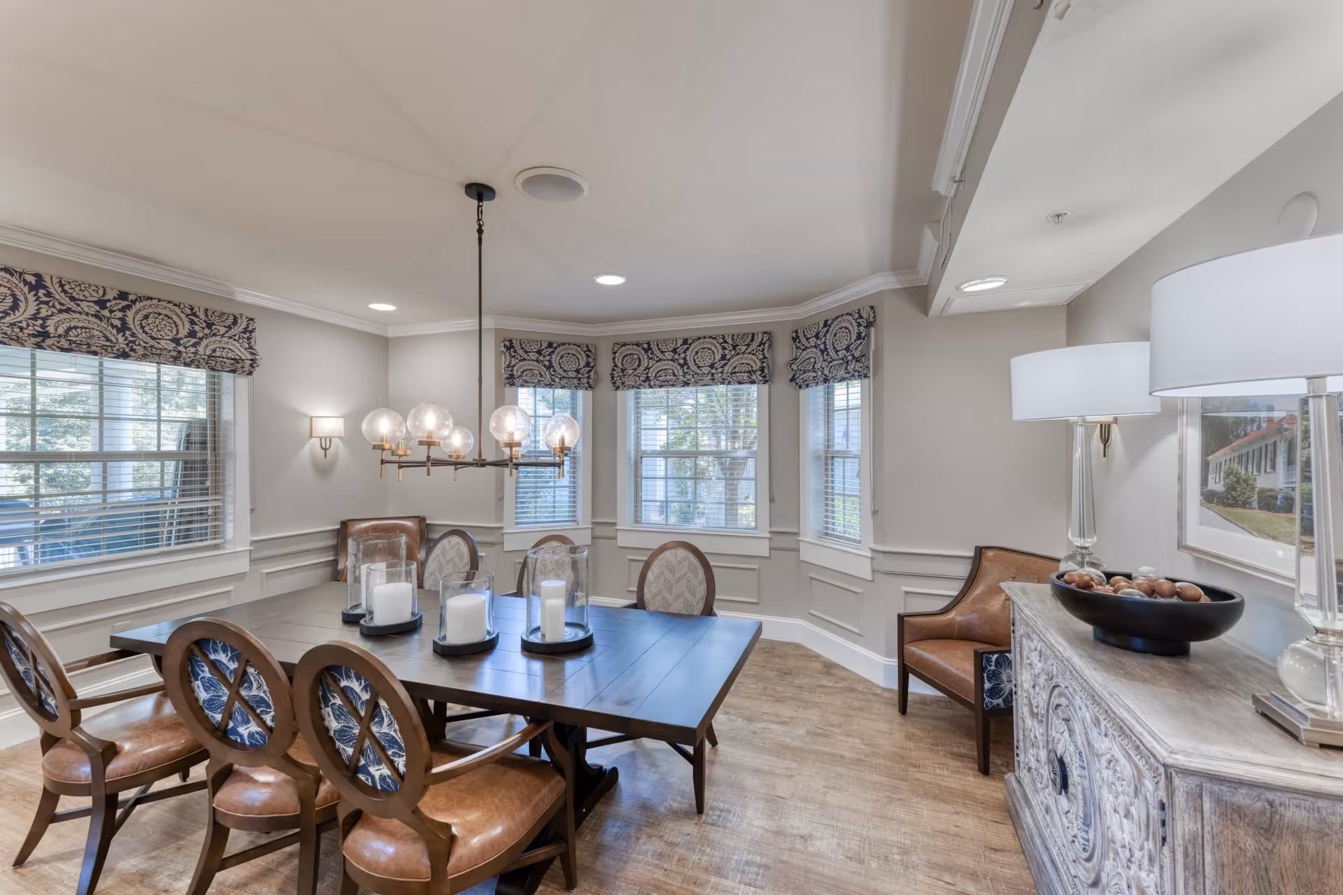 A well-lit dining room with a large wooden table surrounded by six chairs with leather seats and patterned backs. The room features a modern chandelier with globe lights hanging above the table, three large windows with patterned valances, a wooden sideboard with decorative items including a bowl of spheres and two lamps, and light-colored walls with wainscoting and wood flooring.