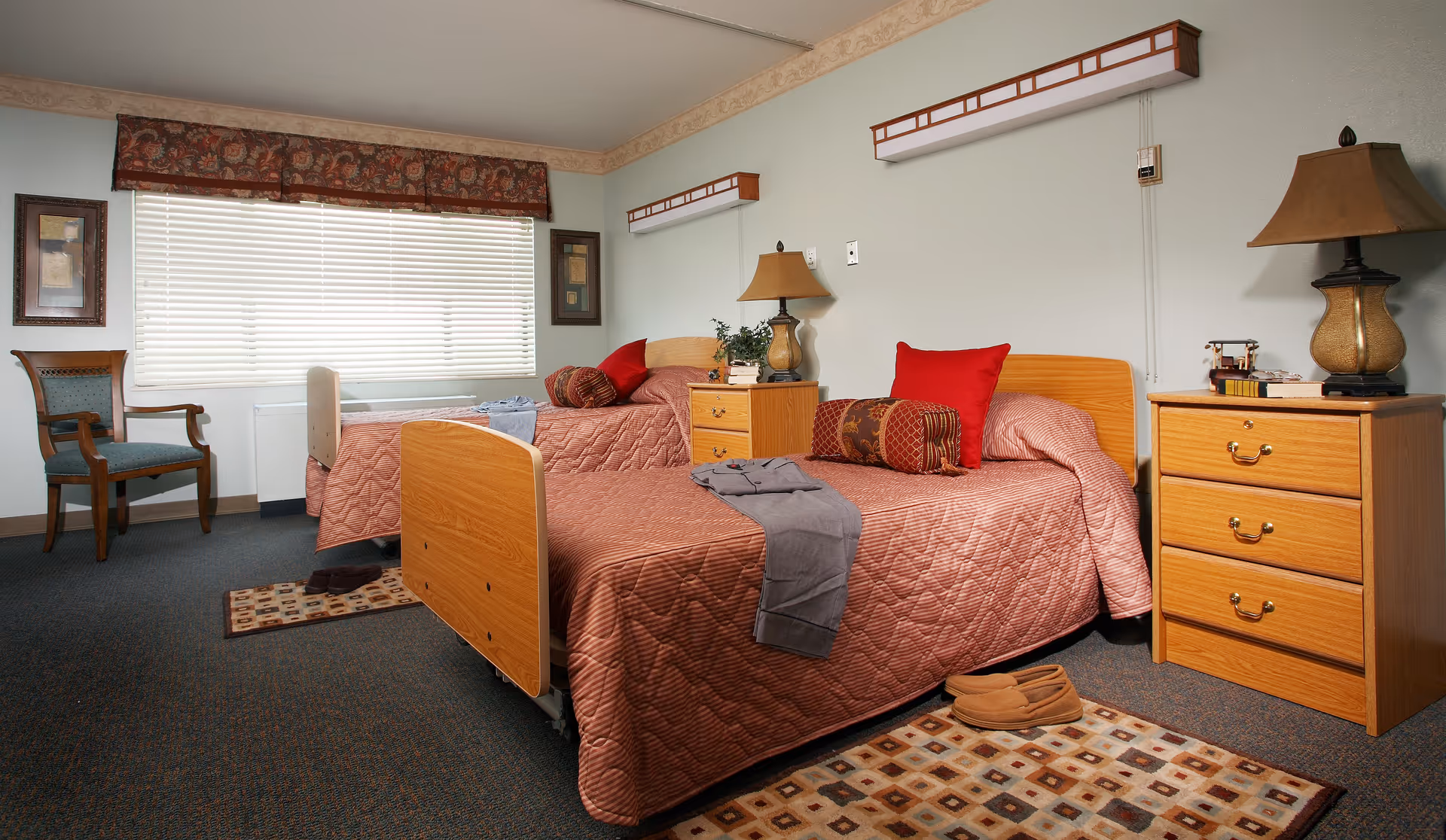 A senior living facility bedroom with two single beds covered in matching quilted bedspreads. Each bed has decorative pillows and a folded blanket. There are two wooden nightstands with lamps, a wooden chair near a window with blinds and a valance, and framed artwork on the walls. The room has carpeted flooring with small area rugs near the beds.