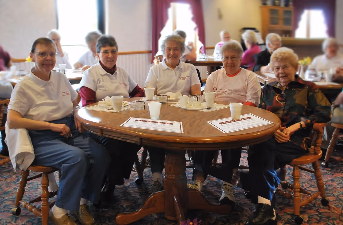 Five elderly women sitting around a wooden round table in a dining area, smiling and enjoying each other's company. The table has disposable cups, plates, and placemats. Other people are visible in the background seated at tables.