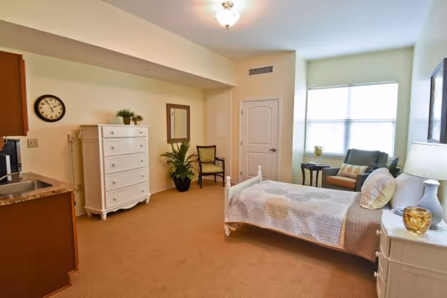 A well-lit bedroom in a senior living facility featuring a bed with a quilted bedspread, a white nightstand with a lamp and decorative items, a comfortable armchair with a side table near a large window, a white chest of drawers, a wall clock, a mirror, a potted plant, and a small kitchenette area with a sink and cabinets.