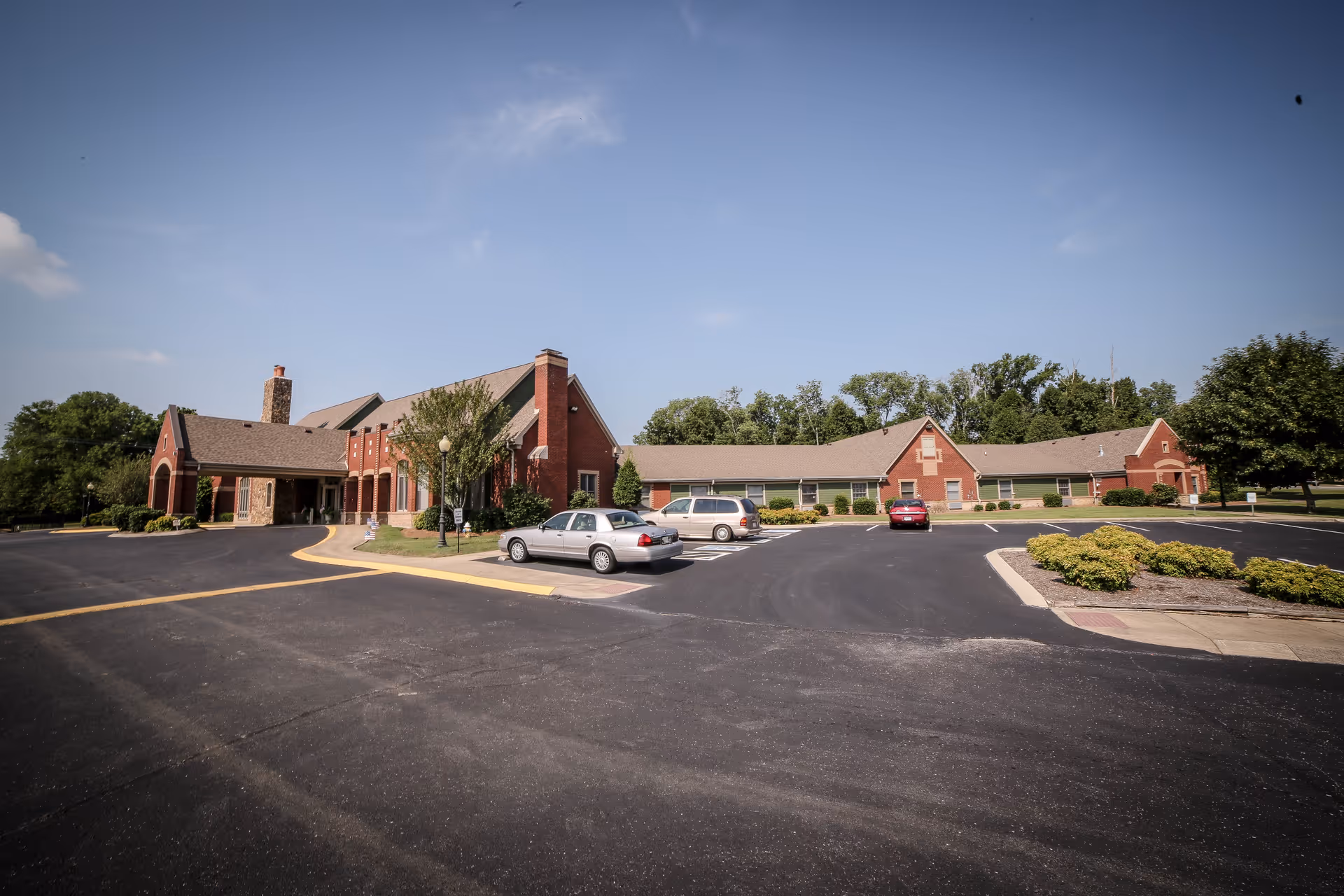 Brick Hearthside Senior Living building front with a parking lot, a few cars, and trees under a blue sky.