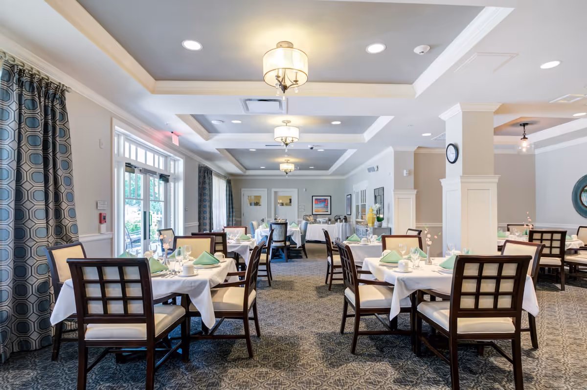 Bright dining room with tables set with white tablecloths, green napkins, and wooden chairs.