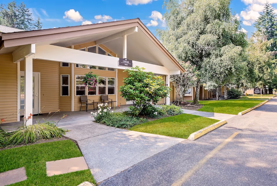 Covered front entrance of a cottage-style senior living building with chairs, hanging planter, shrubs and an adjoining driveway.
