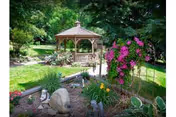 A wooden gazebo sits in a lush garden with a stone path, flowering plants, and a pink-flowered trellis arch.