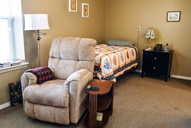A cozy assisted living bedroom featuring a single bed with a colorful quilt, a beige recliner chair with a red plaid blanket draped over one arm, a wooden side table with a remote control, a black nightstand with a vintage-style lamp, telephone, and clock, and a window letting in natural light.