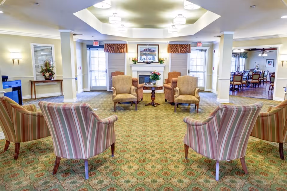 A spacious, well-lit assisted living facility common area with patterned carpet and a seating arrangement of six armchairs around a small round table with a flower vase. The room features a fireplace with a framed picture above it, two exit doors with windows, and a dining area visible in the background to the right.