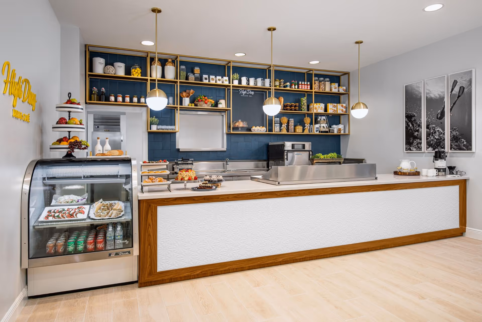 A modern food service counter area with a refrigerated display case containing beverages and desserts on the left, a long counter with baked goods and a salad bar, and open shelving on the back wall holding jars, cups, and food items. Three pendant lights hang from the ceiling, and there are black and white photos on the right wall.