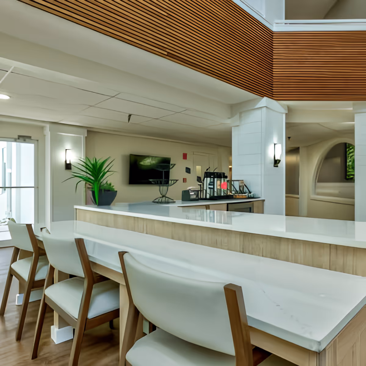 Interior view of a modern senior living facility lounge area with a long white marble countertop and wooden chairs with white cushions. The space features wooden slatted ceiling accents, a potted plant on the counter, wall-mounted lights, and a flat-screen TV mounted on the wall.