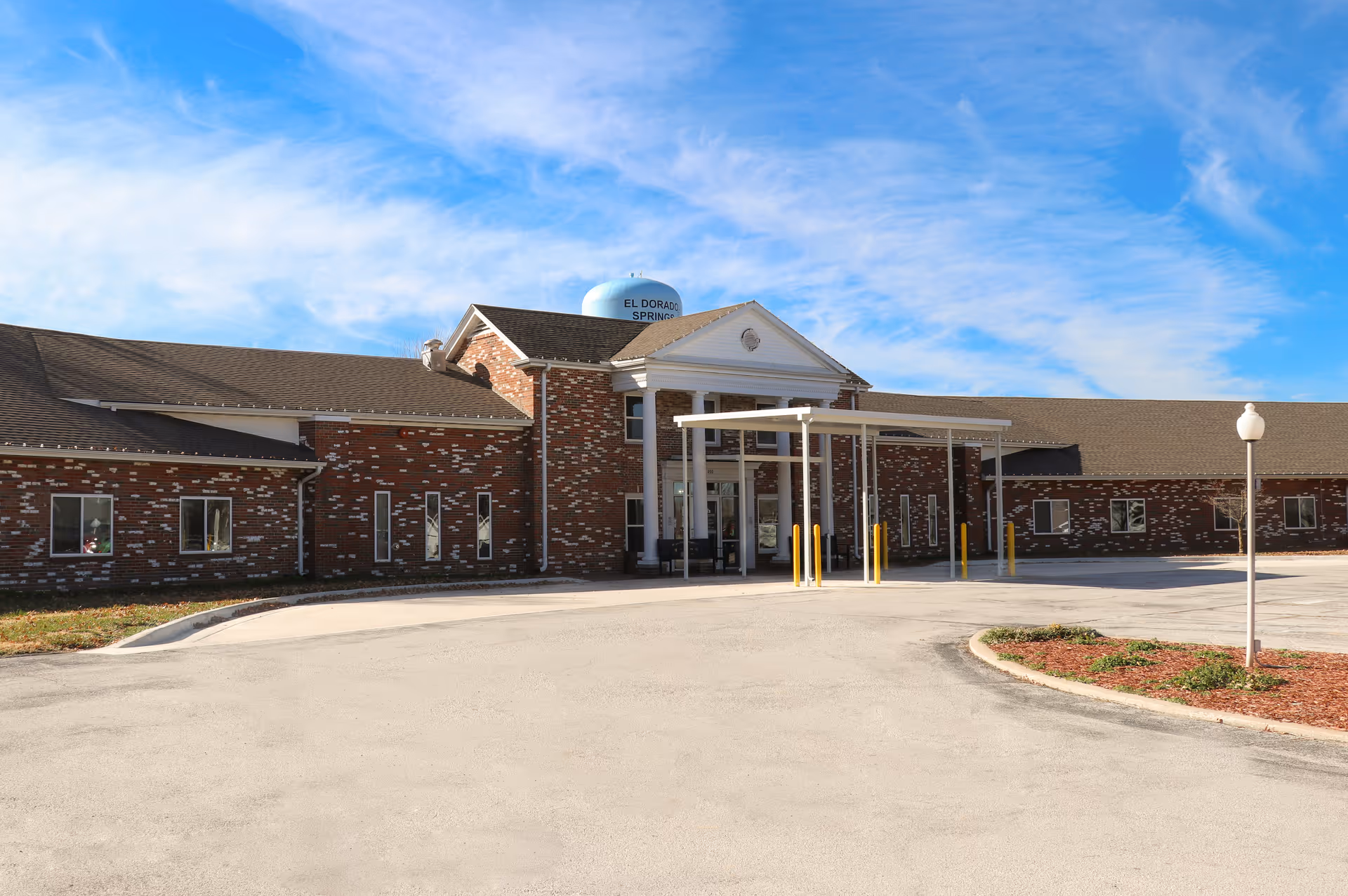 Front exterior view of a single-story brick healthcare facility building with a covered entrance and a circular driveway under a blue sky with light clouds.