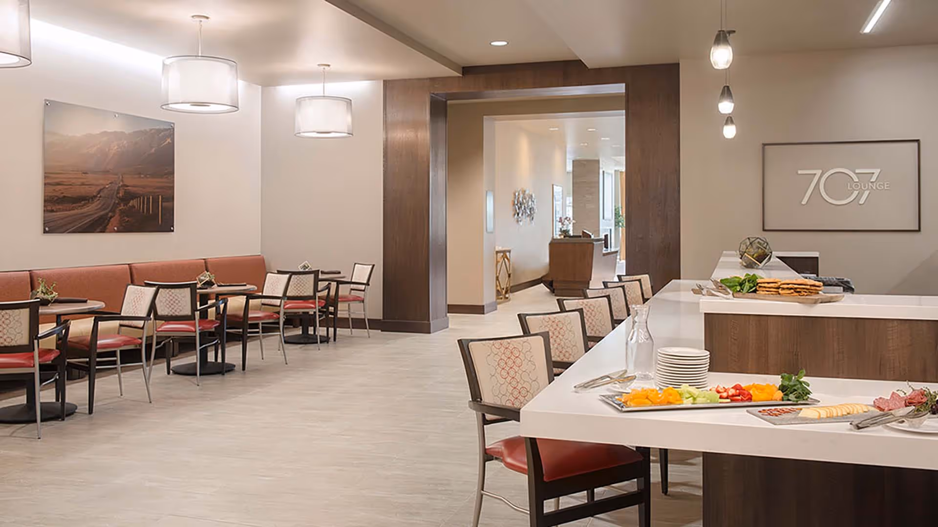Bright modern dining/lounge area with tables and chairs, a long counter with food platters, pendant lights, and framed wall art.