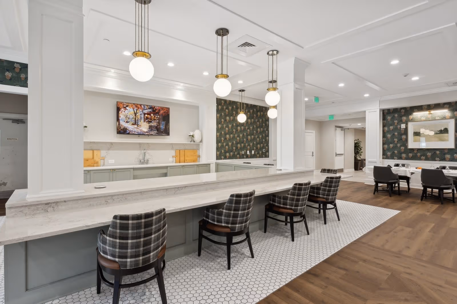 Interior view of a senior living facility dining area featuring a long marble countertop with five plaid upholstered chairs. The space includes hanging globe pendant lights, a wall-mounted TV above a countertop with a sink, and a seating area with tables and chairs in the background. The room has a mix of white and dark green patterned walls and wood flooring.