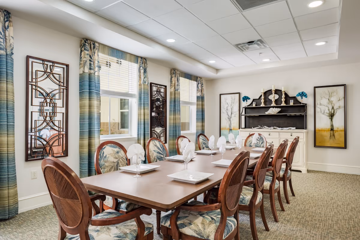 Well-lit dining room with a long table set for ten, upholstered chairs, patterned curtains and wall art.