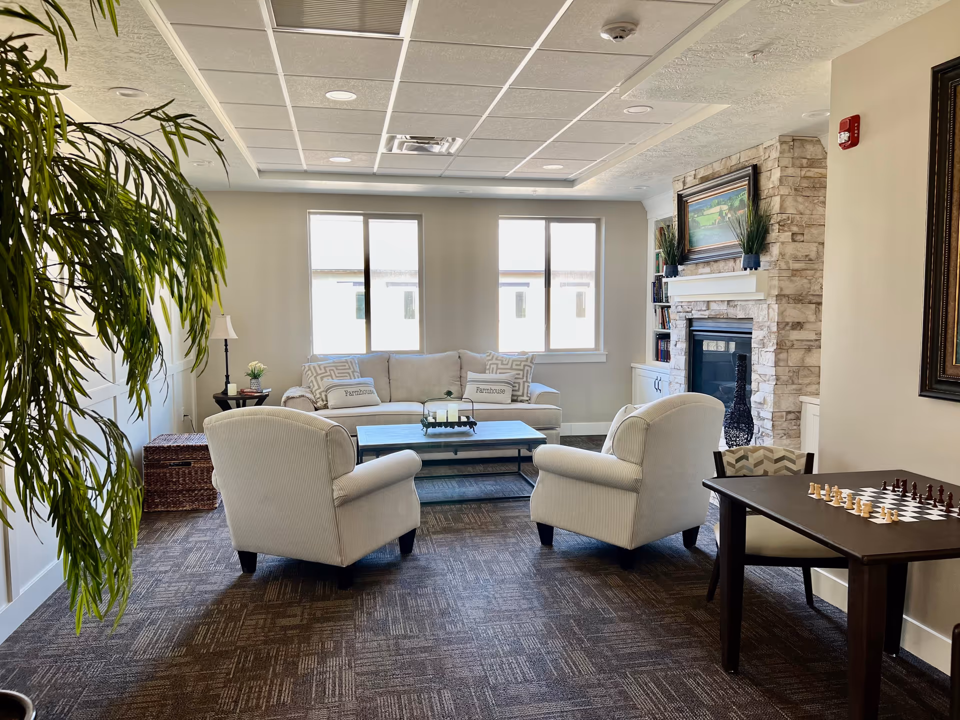A cozy senior living common area with two beige armchairs and a matching sofa arranged around a coffee table. The room features a stone fireplace with a framed painting above it, built-in bookshelves, and large windows letting in natural light. A small table with a chessboard and chairs is positioned to the right, and a large green plant is visible on the left side of the image.